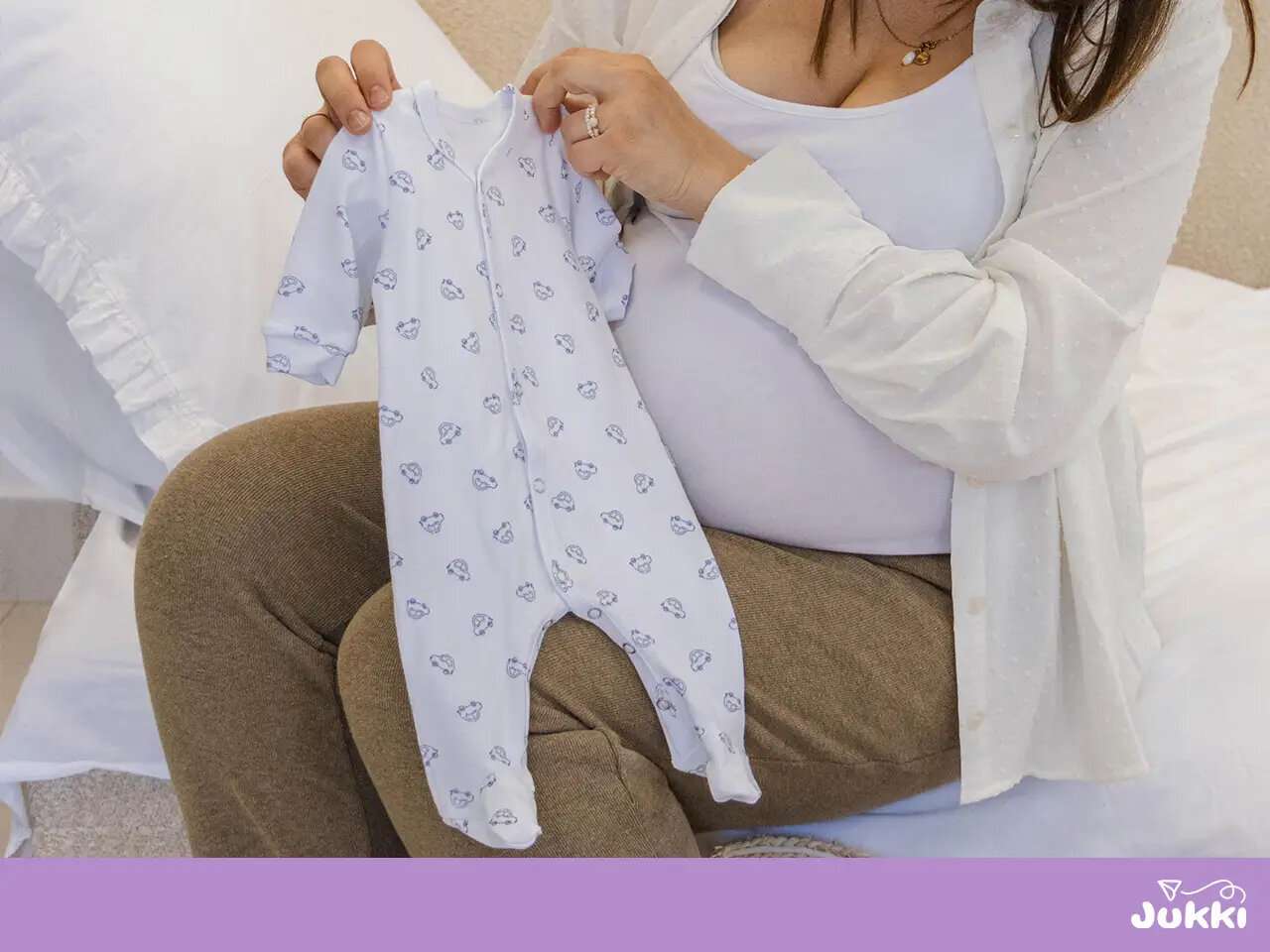 Várandós nő kezében egy fehér, mintás használt babaruha, amelyet adományozásra vagy eladásra szán