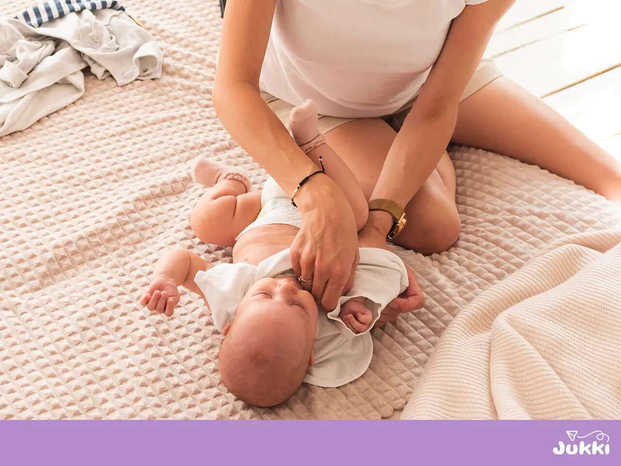 Egy anyuka éppen átöltözteti a babáját a puha ágyon, miközben mellette kinőtt ruhák láthatók